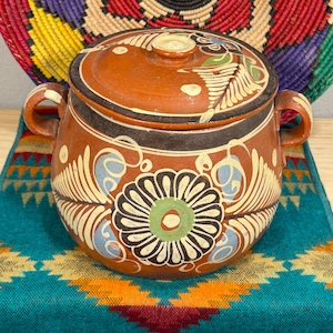 May include: A handcrafted, terracotta-colored ceramic pot with a lid, featuring hand-painted floral designs in white, blue, green, and black. The pot has two handles and sits on a teal textile with an orange and yellow geometric pattern.
