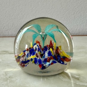 May include: A clear glass paperweight with a colorful interior design. The paperweight features a turquoise floral design at the top and a base of red, yellow, blue, and pink glass fragments. The paperweight is round and sits on a white surface.