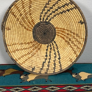 May include: A round, shallow woven basket featuring a coiled design with natural tan and dark brown fibers. The center is a solid dark brown circle, with radiating geometric patterns in dark brown extending toward the braided outer rim.