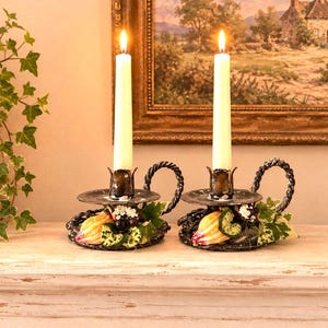 May include: A pair of black metal candle holders with twisted handles, each featuring a lit white taper candle. The base of each holder is decorated with colorful artificial fruit, including small pumpkins and grapes, and green leaves.