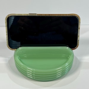 May include: A smartphone in a clear case sits on a light green, semi-circular dish. The dish has a ribbed design and a glossy finish. The phone screen is black. The dish is approximately 15 cm wide.