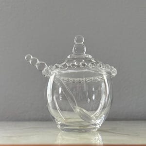 Könnte beinhalten: Eine transparente Zuckerdose aus Glas mit Deckel und Löffel. Die Schüssel ist rund mit einem Perlenrand und einem kleinen Griff am Deckel. Der Löffel liegt in der Schüssel. Das Glas ist transparent.
