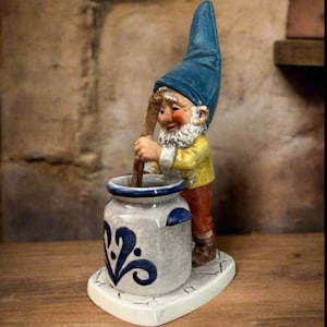 May include: A whimsical garden gnome figurine stirring a pot. The gnome wears a blue hat, yellow jacket, and red pants. The pot is gray with blue floral designs and sits on a white base. The figurine is on a wooden surface.