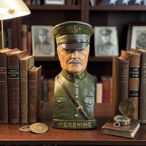 May include: A bust of General Pershing in a military uniform. The olive green bust has a detailed face with a mustache and a military cap. The word "PERSHING" is at the base. The bust is displayed on a shelf with books and a compass.