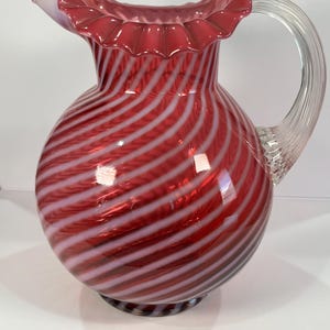 May include: A red and white swirled glass pitcher with a ruffled rim and a clear glass handle. The pitcher has a bulbous body and a round base. The design features alternating red and white stripes.