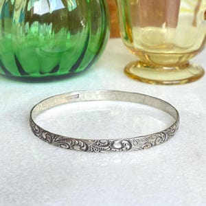 May include: A thin sterling silver bangle bracelet featuring an intricate, embossed floral scrollwork pattern along the exterior surface. The interior of the band is stamped with the text BANCROFT STERLING, indicating its metal composition.