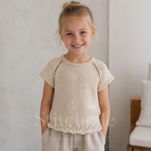 Peut inclure: Un haut en tricot couleur crème à manches courtes, doté d'une encolure ronde, de manches raglan avec des détails ajourés le long des coutures et d'un motif délicat en dentelle festonnée sur l'ourlet inférieur, porté avec un pantalon clair.