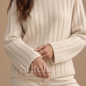 May include: A cream-coloured long-sleeve jumper featuring a vertical ribbed knit pattern, a high neckline, and wide sleeves. The garment is crafted with a consistent texture throughout, showcasing a clean design with a hemmed waist and cuffs.