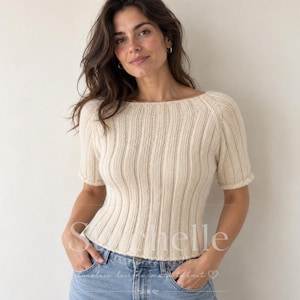 May include: A cream-colored, short-sleeved knit sweater featuring a vertical ribbed texture, a wide boat neckline, and fitted cuffs. The garment is worn with light blue denim jeans. The text "Shelle" is visible across the midsection.
