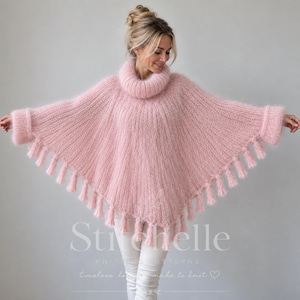 Peut inclure: Un poncho tricoté en maille duveteuse rose clair, doté d'un col roulé haut, de manches longues avec de larges poignets et d'un ourlet orné de pompons pendants. Le vêtement présente un aspect doux et texturé, porté sur un pantalon blanc.