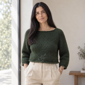 May include: A dark forest green long-sleeved jumper with a wide boat neckline and a chunky knit texture. The jumper features a cropped length and raglan sleeves, paired with light beige high-waisted trousers that have front pleats.