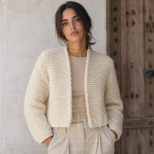 Op de afbeelding: Een crèmekleurig, aan de voorzijde open gebreid vest met een gestructureerd, grof breipatroon. Het kledingstuk heeft lange mouwen en een rechte zoom, gedragen over een bijpassende beige top en een lichte broek, wat een neutraal ensemble vormt.
