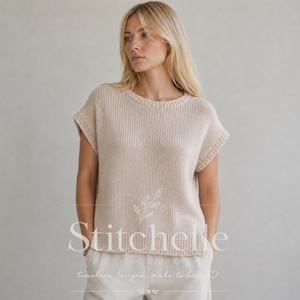 Può includere: Un top in maglia beige a maniche corte con scollo rotondo e bordi a coste su colletto, maniche e orlo. Il capo presenta un motivo a maglia testurizzato ed è indossato sopra pantaloni chiari. Il testo "Stitchelle" è visibile sul davanti.
