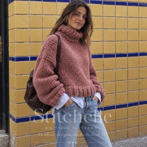 Pode incluir: Um suéter de malha grossa em rosa empoeirado com gola alta, mangas compridas e bainha canelada. O suéter é usado sobre uma camisa branca com colarinho e combinado com jeans azul claro. Uma mochila de couro marrom escuro é usada em um ombro.