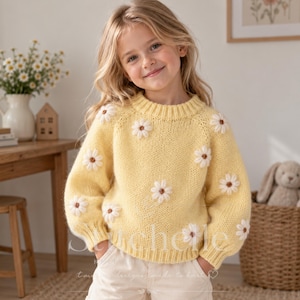 Può includere: Un maglione in maglia a maniche lunghe color giallo pallido con scollo rotondo e polsini a coste. Il maglione è decorato con diverse applicazioni a forma di margherita bianca, ognuna con un piccolo centro marrone, sparse sul davanti e sulle maniche.
