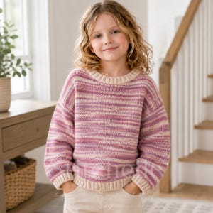 May include: A long-sleeved knit jumper featuring a variegated horizontal stripe pattern in shades of pink, purple, and cream. The garment has a round neckline and ribbed cuffs and hem in a solid cream colour, worn with light-coloured trousers.