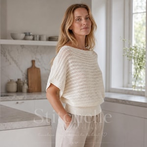 Op de afbeelding: Een gebreide top in off-white met korte mouwen, een brede boothals, een luchtig opengewerkt breipatroon op het lijfje en een effen boord met ribbelsteek in de taille. Het kledingstuk wordt gecombineerd met een lichte, casual gebreide broek.