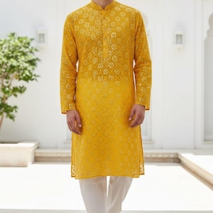 May include: A man wearing a yellow kurta with intricate floral embroidery, paired with white pants. The long-sleeved tunic has a mandarin collar. The text "WEDDING KURTA" is visible at the bottom left of the image.