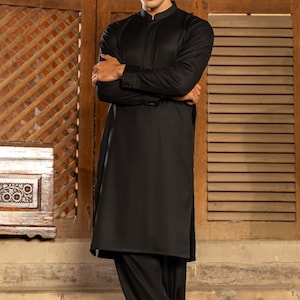 May include: A man wearing a black long-sleeved shirt and black pants. He is standing in front of a wooden wall with a lattice pattern.