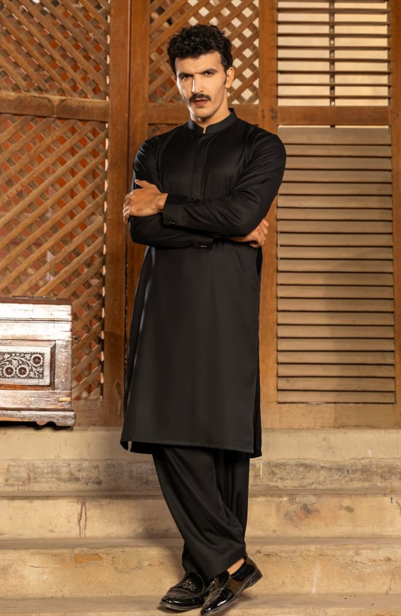 May include: A man wearing a black long-sleeved shirt and black pants. He is standing in front of a wooden wall with a lattice pattern.