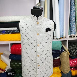 May include: A white, patterned wedding kurta vest with a mandarin collar and wooden buttons. The vest has a black pocket square and is displayed on a mannequin. The text "WEDDING KURTA" is visible.