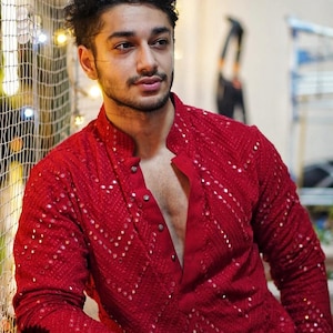 Puede incluir: Un kurta rojo con cuello mandarín y mangas largas. El kurta está adornado con pequeños detalles reflectantes dispuestos en un patrón diagonal. La imagen incluye el texto "WEDDING KURTA" en un logotipo circular.