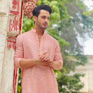 Puede incluir: Un hombre lleva una kurta rosa con un diseño de espejo plateado. La kurta tiene un cuello mandarín y un cierre de botón. El hombre está de pie frente a un pilar blanco con acentos rojos.