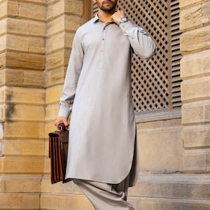 May include: A man wearing a light gray long-sleeved shirt and matching pants. He is holding a brown leather briefcase and is standing in front of a building with a wooden lattice window.