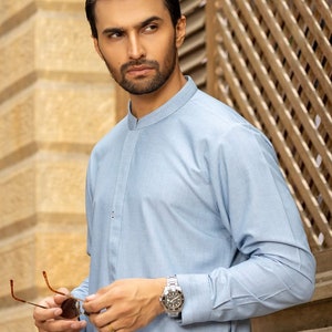 May include: A man wearing a light blue collared shirt with a mandarin collar. He is wearing a silver watch on his left wrist and holding a pair of brown sunglasses.
