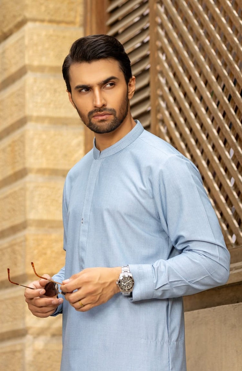 May include: A man wearing a light blue collared shirt with a mandarin collar. He is wearing a silver watch on his left wrist and holding a pair of brown sunglasses.