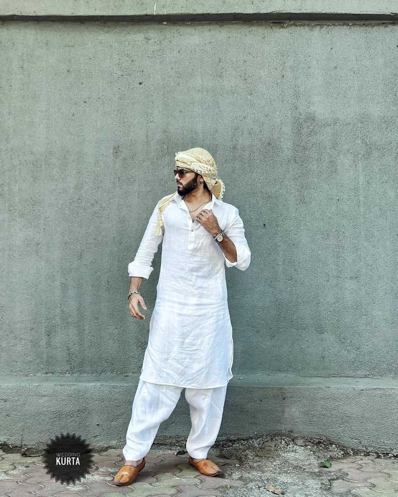 May include: A man wearing a white long-sleeved kurta with a white turban. The kurta has a button-up front and a long, flowing skirt. The man is standing in front of a gray wall.