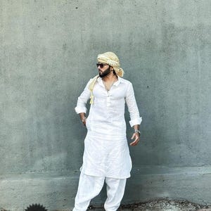 May include: A white long-sleeved kurta with a button-up collar and a matching white shalwar. The man is wearing a gold turban and sunglasses. The text "Wedding Kurta" is visible in the lower left corner of the image.