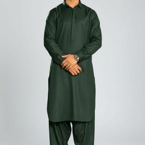 May include: A dark green kurta suit, featuring a long-sleeved shirt with a collared neckline and matching loose-fitting trousers. The outfit is paired with brown dress shoes. The text "WEDDING KURTA" is visible at the bottom left.