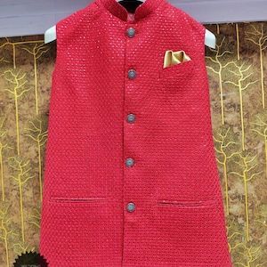 Könnte beinhalten: Eine rote, ärmellose Hochzeits-Kurta mit strukturiertem Muster. Die Kurta hat einen Stehkragen, dekorative Knöpfe und eine Tasche mit goldenem Akzent. Das Kleidungsstück wird auf einem weißen Kleiderbügel präsentiert. Der Text "Wedding Kurta" ist sichtbar.