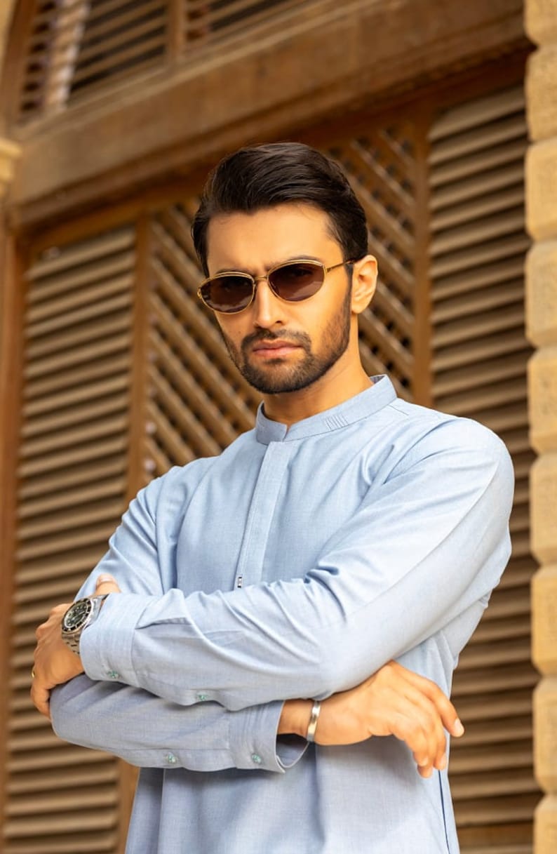 May include: A man wearing a light blue collared shirt with a mandarin collar, standing in front of a wooden slat wall. He is wearing brown sunglasses and a silver watch.