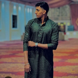 Puede incluir: Una kurta de color verde oscuro con estampado y cierre de botones. La kurta la lleva puesta una persona de pie en una habitación con una alfombra de color rojo y dorado con estampado.