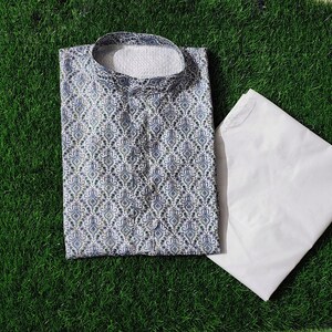 May include: A light blue and white patterned kurta shirt with a matching white pajama set, displayed on a green surface. The kurta features a mandarin collar and intricate design. The word "KURTA" is printed on a black circle.