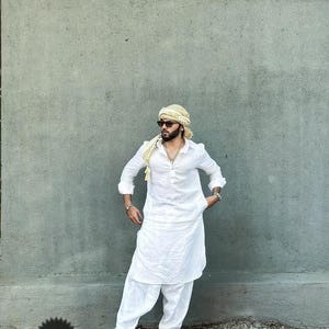 May include: A man wearing a white kurta with a long, flowing design. He is wearing a yellow turban and sunglasses. The kurta has a button-up front and long sleeves. The man is standing in front of a gray wall.