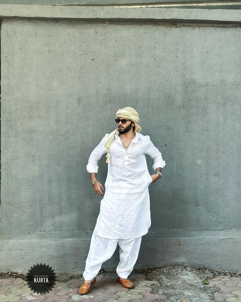 May include: A man wearing a white kurta with a long, flowing design. He is wearing a yellow turban and sunglasses. The kurta has a button-up front and long sleeves. The man is standing in front of a gray wall.