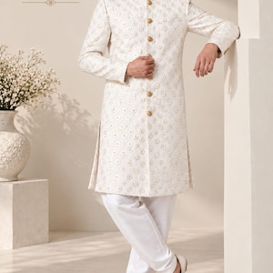 May include: An off-white wedding kurta featuring a repeating circular gold embroidered pattern, a mandarin collar, and a front button placket with gold buttons. The garment is paired with plain white trousers and light-colored pointed shoes.