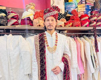Puede incluir: Una kurta blanca con bordados intrincados, combinada con un chal de terciopelo granate y un collar de oro. El hombre lleva un tocado tradicional indio.