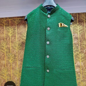 May include: Emerald green vest with a textured pattern, featuring a mandarin collar and four decorative buttons. A gold pocket square accents the chest pocket. The vest is displayed on a hanger against a patterned background. The text "WEDDING KURTA" is in the lower left corner.