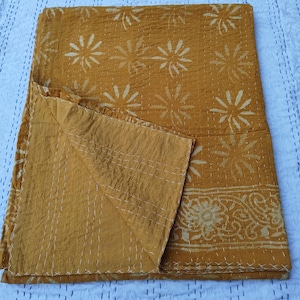May include: Mustard-colored quilt with a floral pattern and white stitching. The quilt features a repeating design of stylized flowers and a decorative border. The fabric appears soft and has a slightly wrinkled texture, suggesting it is made of cotton or a similar material.