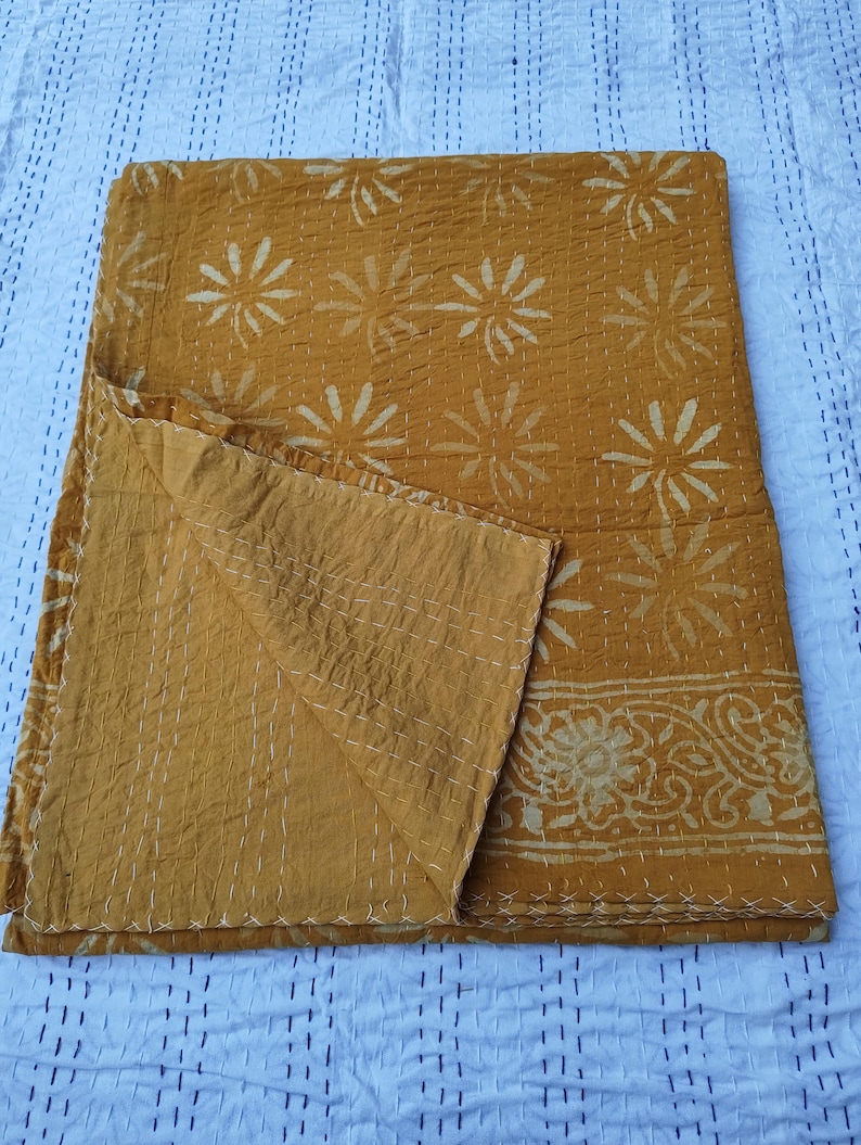 May include: Mustard-colored quilt with a floral pattern and white stitching. The quilt features a repeating design of stylized flowers and a decorative border. The fabric appears soft and has a slightly wrinkled texture, suggesting it is made of cotton or a similar material.