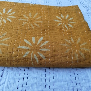 May include: Mustard yellow quilt with large, white floral designs. The quilt features white stitching throughout, creating a textured appearance. The background is a white fabric with a dark blue stitched pattern.