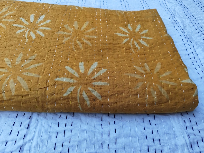 May include: Mustard yellow quilt with large, white floral designs. The quilt features white stitching throughout, creating a textured appearance. The background is a white fabric with a dark blue stitched pattern.