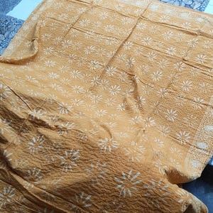 May include: Mustard-colored textile with a repeating white floral pattern. The fabric is textured and appears to be folded, with the pattern visible across the surface. The edges have a decorative border. The textile is likely a blanket or throw.