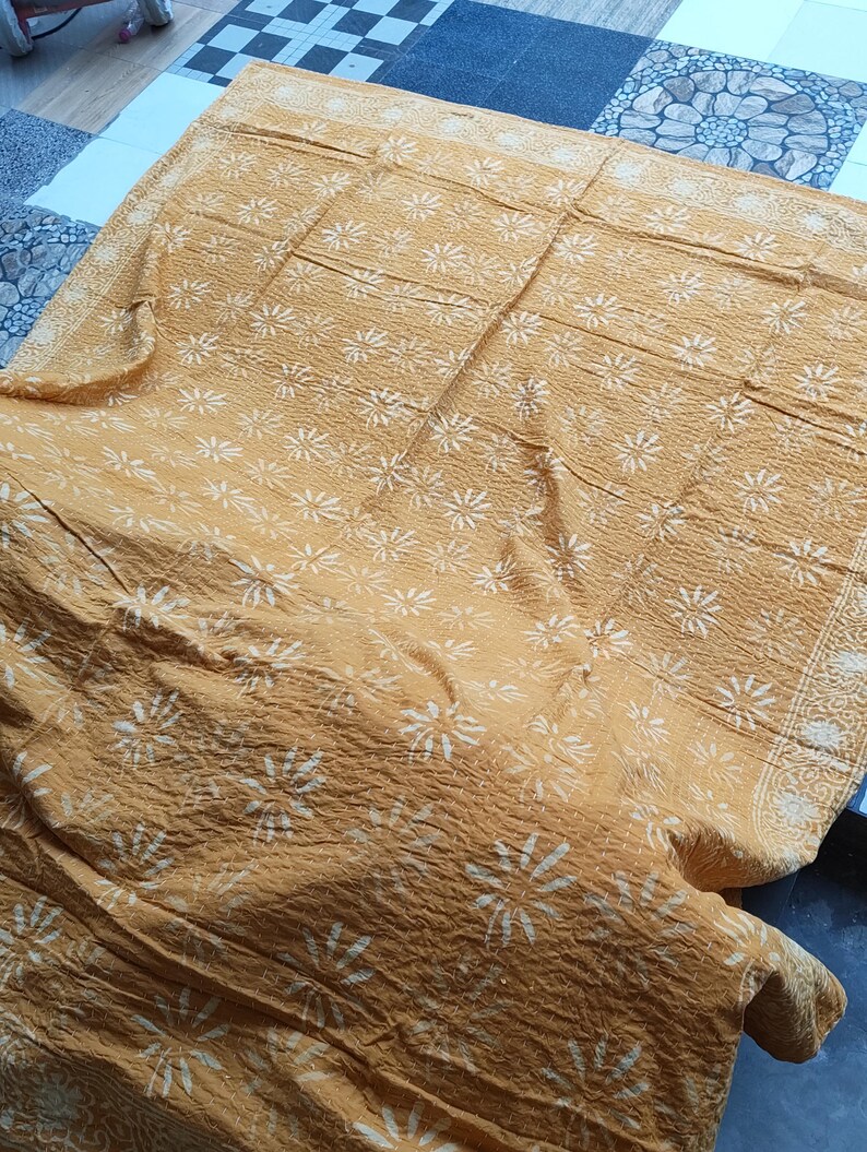 May include: Mustard-colored textile with a repeating white floral pattern. The fabric is textured and appears to be folded, with the pattern visible across the surface. The edges have a decorative border. The textile is likely a blanket or throw.