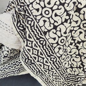 May include: A folded, cream-colored quilt with a repeating black pattern. The design features swirling shapes and geometric borders. The quilt is made of a soft material and has visible stitching.