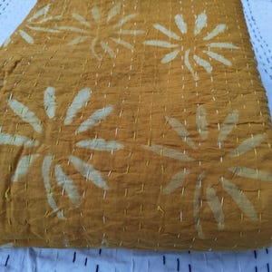 May include: Folded mustard-colored textile with large, white floral patterns. The fabric is stitched with white thread, creating a textured appearance. The textile is resting on a white surface with a blue and black striped pattern.